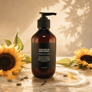 A bottle of Francis Michael Hydrating Lotion stands on a reflective surface with a white cream swirl around it, surrounded by sunflowers and leaf shadows in warm natural light.