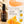 Francis Michael Nourishing Serum bottle on a wooden surface with orange slices, daisies, and a peach slice graphic on a beige background with the text 'brightens' and product benefits.