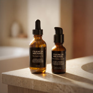 Two bottles of Francis Michael skincare products, Revitalizing Oil and Nourishing Serum, on a stone surface with a blurred background.