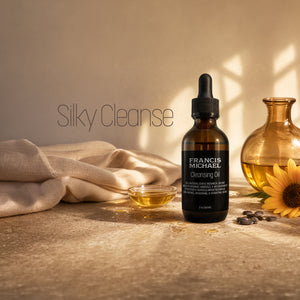 Francis Michael 'Silky Cleanse' Cleansing Oil in an amber glass dropper bottle, styled on a stone surface with a beige cloth, a small dish of oil, coffee beans, and a yellow sunflower under warm, dappled sunlight.